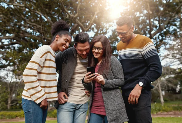 Grupo de jóvenes amigos multirraciales de pie juntos mirando el teléfono móvil en el parque sonriendo - caliente al aire libre —  Fotos de Stock