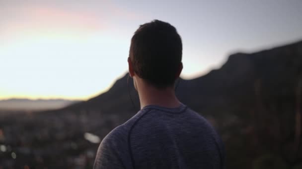 Close-up van sportieve jonge man luisteren muziek op oortelefoons kijken naar het stadsgezicht uitzicht vanaf Mountain Top-inspiratie — Stockvideo