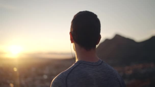Vista lateral de un hombre de fitness escuchando música usando auriculares con vistas a la vista del paisaje urbano en la madrugada - admirando la naturaleza — Vídeo de stock