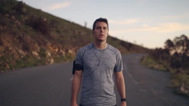 Retrato de un joven atleta confiado que usa auriculares escuchando música mientras corre por un camino sinuoso en la montaña, corriendo en la naturaleza — Vídeo de stock