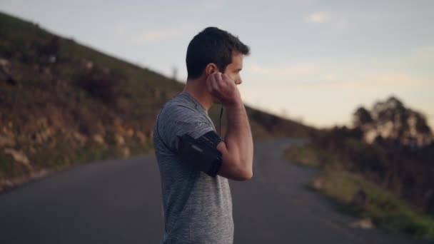 Genç sportif adam dağ yolda kulaklık ile müzik dinlemek için kol bandı üzerinde bir akıllı telefon kullanarak - motivasyon ve kararlılık — Stok video