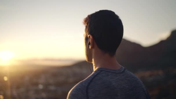 Boční pohled na mužského sportovce, který nosí sluchátka užívající ranní sluneční světlo a výhled na cityy z horského muže, který si užívá východu slunce a přírody — Stock video