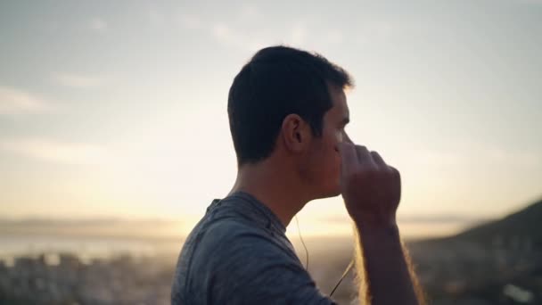 Schöner junger Mann, der seine Kopfhörer einsetzt, bevor er joggt, lächelt und in die Kamera blickt, die gegen den morgendlichen Sonnenaufgang steht — Stockvideo