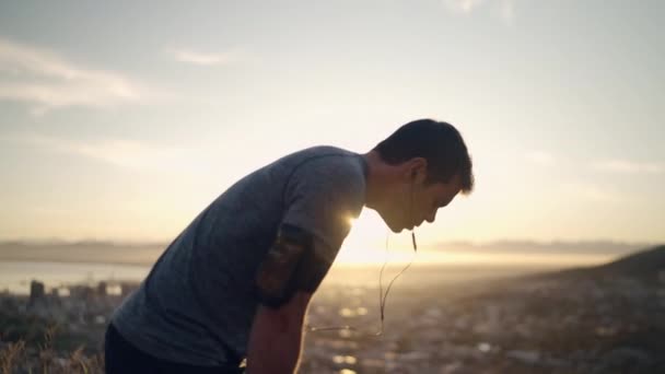 A tired male runner taking a rest after running hard in morning sunlight on top of the mountain - wide shot of an exhausted man runner — Stock Video