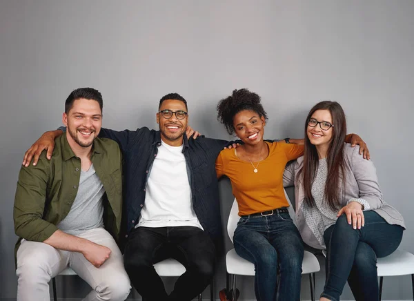 Ritratto di giovani amici multietnici seduti insieme felicemente con le braccia intorno a vicenda sulla sedia sorridente e guardando la macchina fotografica — Foto Stock