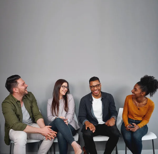 Jóvenes amigos multirraciales están sentados alegres en una fila en la silla blanca divirtiéndose juntos contra la pared gris — Foto de Stock