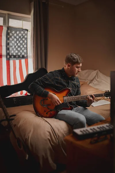 Giovane musicista in abiti casual seduto da solo in camera da letto trascorrere del tempo a suonare la chitarra a casa — Foto Stock