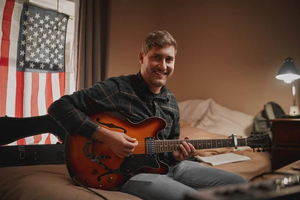 Ritratto di un giovane musicista sorridente che suona la chitarra nella sua camera da letto di notte guardando la macchina fotografica — Foto Stock