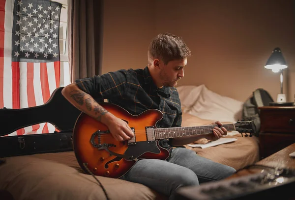 Jeune homme concentré en tenue décontractée assis sur le lit jouant de la guitare le soir — Photo