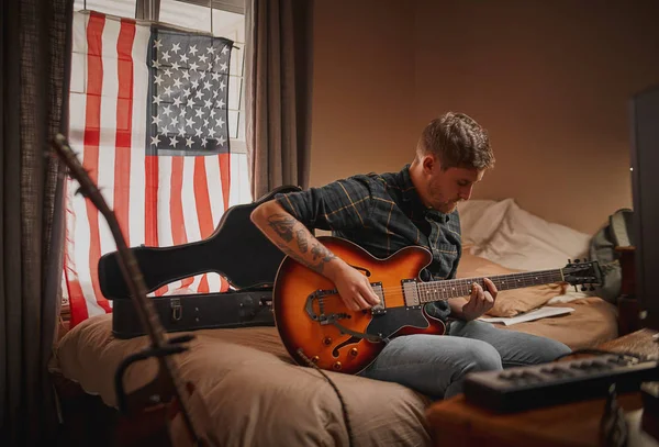 Młody gitarzysta człowiek w domu siedzi na łóżku przed oknem z usa flag gra na gitarze — Zdjęcie stockowe