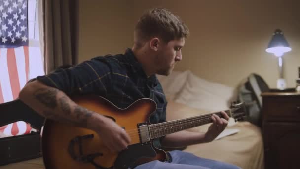 Närbild av en erfaren konstnärlig ung man som sitter på sängen och spelar gitarr efter sin passion — Stockvideo