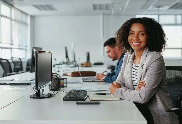 Succesvolle glimlachende jonge zakenvrouw met haar armen gekruist kijkend naar camera en mannelijke collega werkt aan een bureau op de achtergrond — Stockfoto