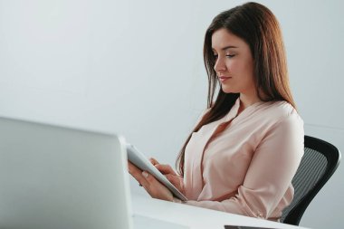 Güzel genç iş kadını ofiste dijital tablet kullanarak dizüstü bilgisayarın önünde oturuyor.
