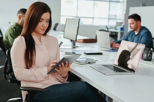 Una atractiva joven mujer de negocios sonriente usando tableta digital con colegas trabajando en el escritorio en segundo plano en la oficina - startup office —  Fotos de Stock