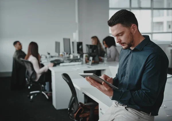 Portret van jonge zakenman leunend op bureau met behulp van tablet pc en zijn collega 's werken op de achtergrond — Stockfoto