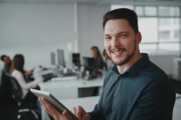 Portret van een zelfverzekerde glimlachende jonge zakenman die digitale tablet in de hand houdt met zijn collega 's die op de achtergrond achter het bureau werken — Stockfoto