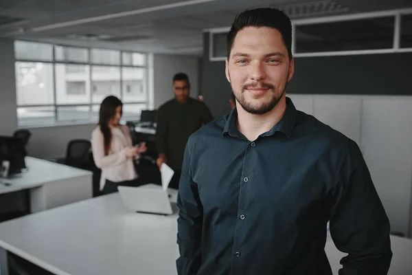 Portret van een professionele jonge zakenman glimlachend en kijkend naar de camera terwijl andere zakenmensen praten op de achtergrond op kantoor — Stockfoto
