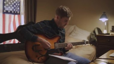 Kendini adamış genç bir adam yatakta oturup gitar çalıyor ve evde notalar yazıyor.