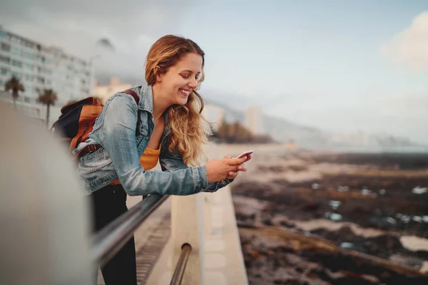Усміхнена блондинка молода жінка мандрівник з рюкзаком спирається на перила на міській вулиці, використовуючи свій мобільний телефон — стокове фото