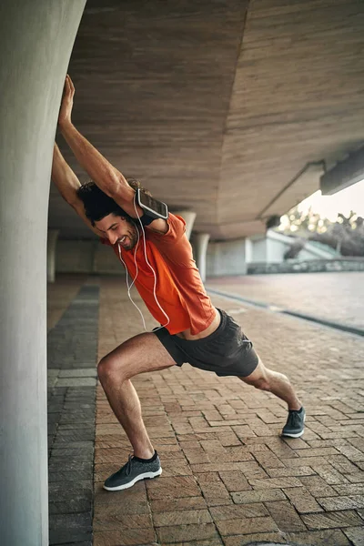 Vista lateral de um alegre fitness jovem corredor masculino com dispositivos eletrônicos esticando a mão na coluna sob a ponte — Fotografia de Stock