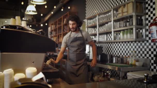 Веселый кавказский владелец кафе в фартуке и чистит кофеварку тряпкой, наклоняясь и стоя за прилавком — стоковое видео