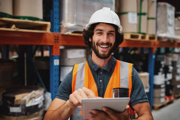 Bello allegro giovane caucasico sorridente supervisore con casco bianco utilizzando tablet digitale per il lavoro mentre in piedi in magazzino contro le merci — Foto Stock