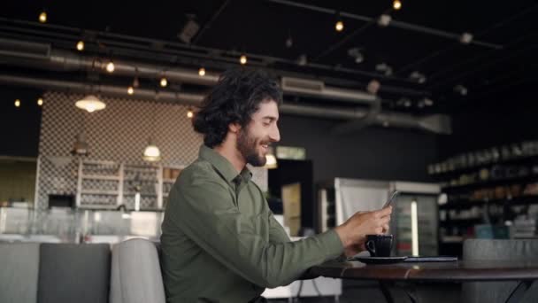 Pemuda tampan tertawa sambil mengirim pesan di smartphone duduk di kafe modern — Stok Video