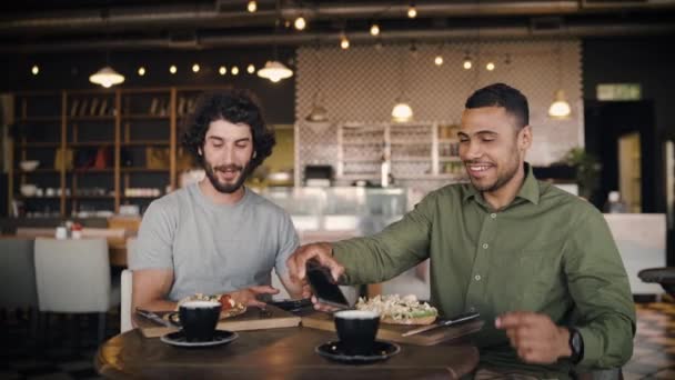 Joven hombre afroamericano con amigo caucásico haciendo clic en la foto con teléfono inteligente de bruschetta italiana con café en la cafetería mientras disfruta del almuerzo en la cafetería moderna — Vídeos de Stock