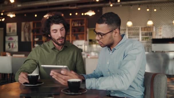 Africký obchodník ukazující služby společnosti zákazníkovi sedícímu v moderní kavárně pomocí digitálního tabletu — Stock video