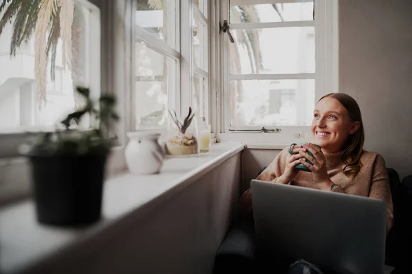 Succesvolle jonge zakenvrouw met laptop thuis tijdens quarantaine met een koffiekopje in de hand terwijl ze uit het raam kijkt — Stockfoto