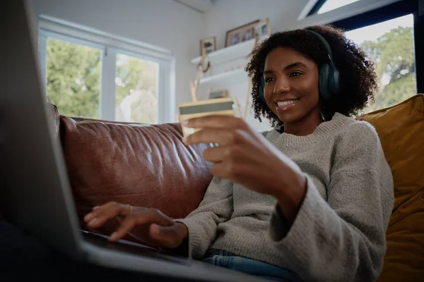 Gelukkige vrouw met draadloze bluetooth hoofdtelefoon en het luisteren naar muziek maken online betaling met behulp van debetkaart en laptop thuis — Stockfoto