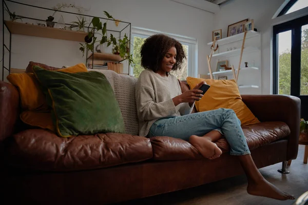 Mujer africana sonriente sentada en un cómodo sofá usando un teléfono inteligente en casa durante el encierro — Foto de Stock