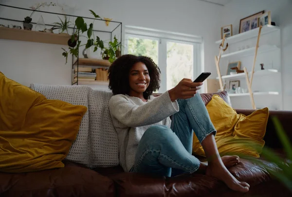 Mujer joven sentada en el interior viendo la televisión y manteniendo el control remoto en el sofá — Foto de Stock