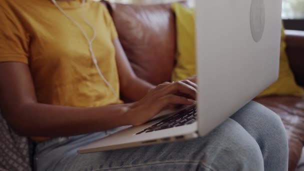 Tilt up shot seorang wanita muda dengan earphone menggunakan laptop-nya duduk di sofa di ruang tamu bekerja dari rumah selama pandemi coronavirus — Stok Video