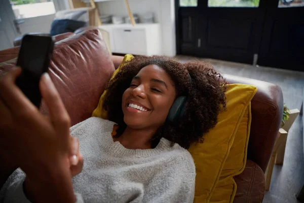 幸せなアフリカの女性は自宅でスマートフォンを使用してソファに休みますヘッドフォンで音楽を聴く — ストック写真