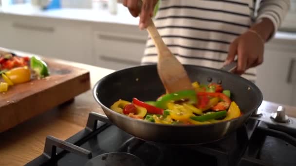 Jovem em casa usa espátula de madeira para atirar fatias coloridas de pimentão na frigideira — Vídeo de Stock
