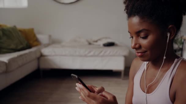 運動中に自宅でスマートフォンにメッセージを送信するイヤホンのアフリカ系アメリカ人女性の笑顔の肖像画 — ストック動画