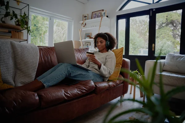 Afro-Amerikaanse jonge vrouw ontspannen op de bank luisteren naar muziek terwijl het maken van online betaling met behulp van bankkaart en laptop — Stockfoto