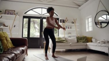 Afro saçlı Afrikalı genç bir kadın evde dizüstü bilgisayarda spor videosu izlerken yoga minderinde egzersiz yapıyor.