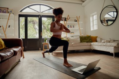 Sağlıklı Afrikalı bir kadın laptopta video izlerken evde yoga minderinde elleriyle egzersiz yapıyor.