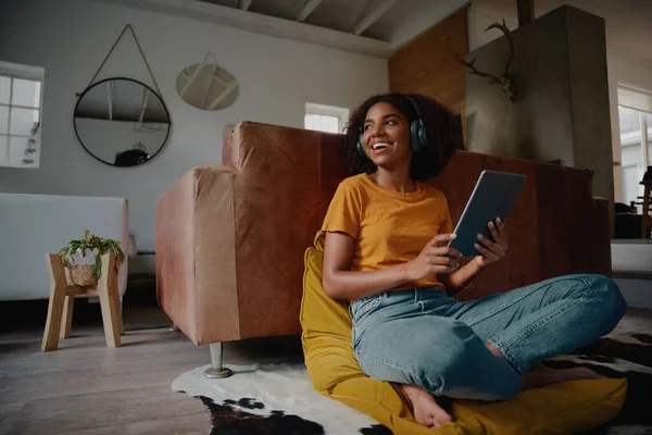 Aantrekkelijke Afrikaanse vrouw thuis zitten op de vloer met behulp van digitale tablet en koptelefoon weg te kijken — Stockfoto