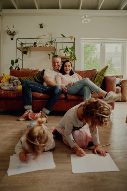 İki çocuk yerde yatıp çizim yaparken ebeveynler onları izlemekten zevk alıyor.