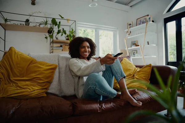Mooie jonge vrouw kijken televisie zitten omringd door kussens op een comfortabele bank met de afstandsbediening in haar hand — Stockfoto
