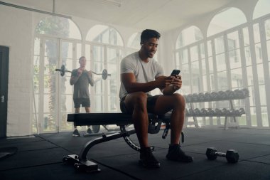 Two multiracial young men wearing sports clothing exercising and using mobile phone at the gym