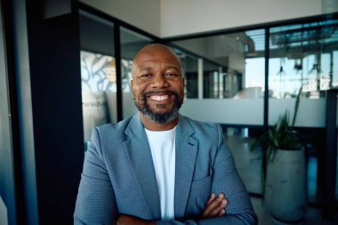 Portrait of smiling African mature businessman in blazer standing arms crossed in modern office
