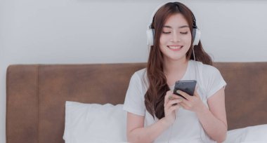 Güzel Asyalı kadınlar kulaklık takıp akıllı telefonlarından müzik dinliyorlar. Beyaz yatak odasındaki yatakta.