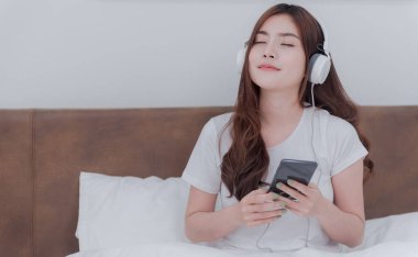 Güzel Asyalı kadınlar kulaklık takıp akıllı telefonlarından müzik dinliyorlar. Beyaz yatak odasındaki yatakta.