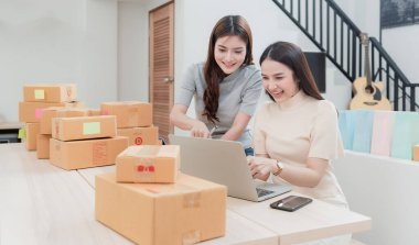İki güzel Asyalı kadın internetten dizüstü bilgisayarların siparişlerini kontrol ediyor. Ve mutlu bir gülümseme ile karton kutu paketleyin, yeni normal bir online iş olmak