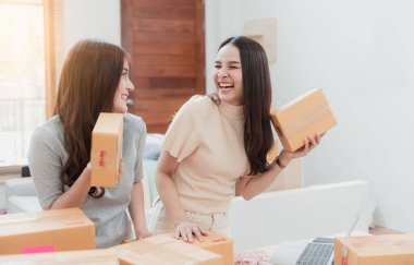 İki güzel Asyalı kadın bir karton kutuyu kaldırıyor. Mutlu bir gülümseme ile, yeni normal bir online iş olmak