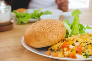 Kahvaltı omletleri, ekmekler, hamburgerler ve sebzeler beyaz bir tabakta, bulanık arka tarafta yenir.